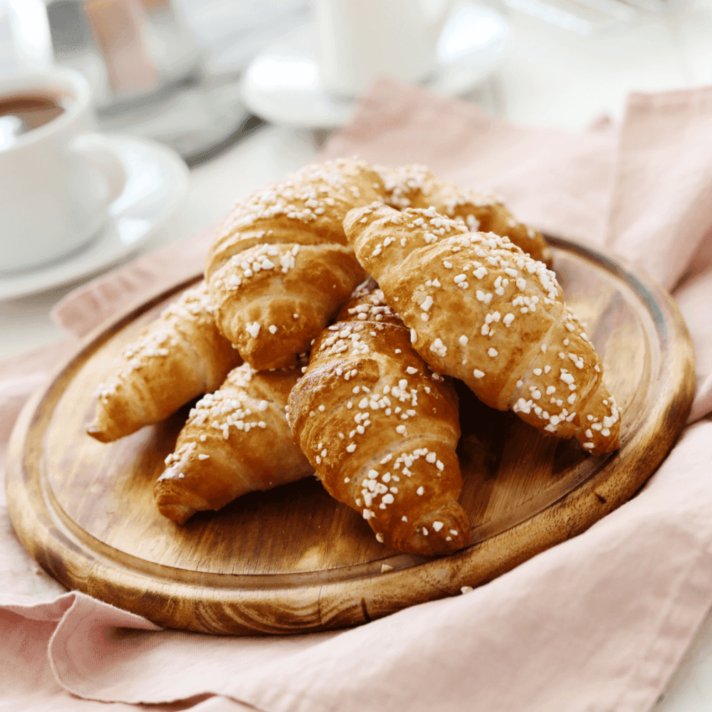 A wooden plate with six sugar-topped croissants on a pink cloth, with a cup of coffee in the background. - Home Instead