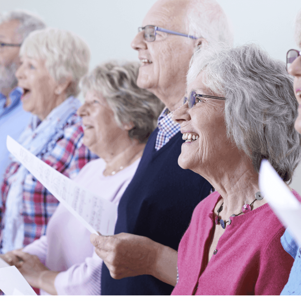 A group of older adults smiling and holding song sheets while singing together. - Home Instead