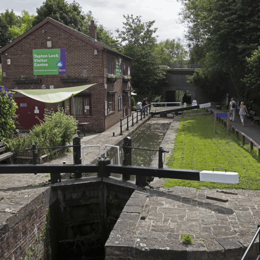 A canal lock with a brick building labeled "Tapton Lock Visitor Centre," surrounded by greenery and a paved path. - Home Instead
