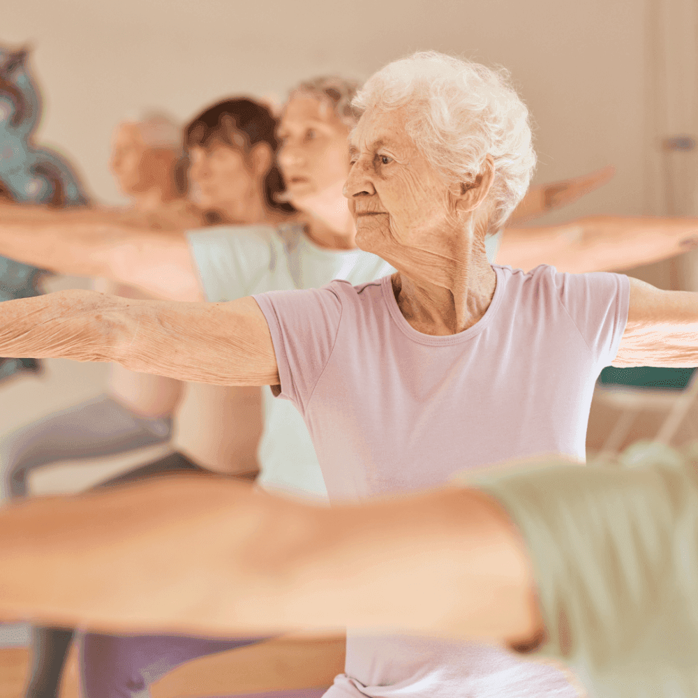 Elderly women practicing yoga together in a studio, focusing on balance with arms extended in a Warrior II pose. - Home Instead
