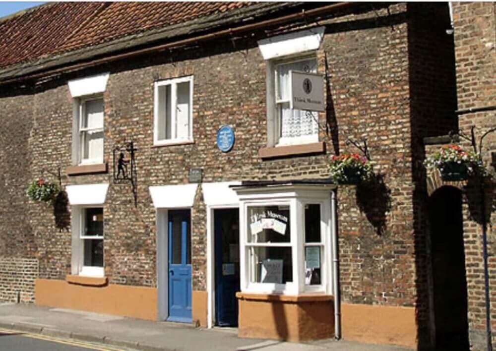 A brick building with a blue door and a "Teale Museum" sign. Flowers hang by the windows, and a blue plaque is on the wall. - Home Instead