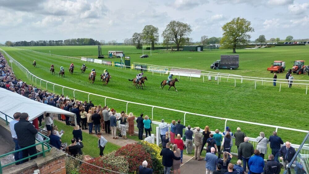 Horse race in progress with an audience watching from the sides of a green race track under a partly cloudy sky. - Home Instead