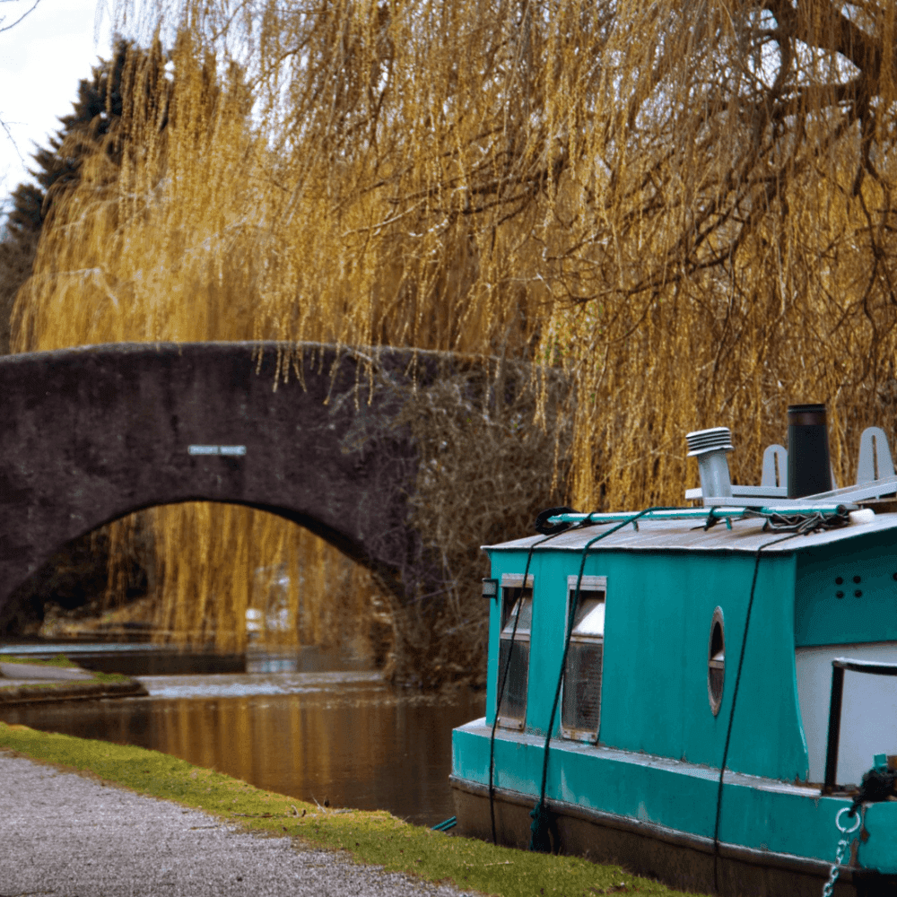 A turquoise canal boat docked near a stone bridge with cascading willow trees by a serene waterway. - Home Instead