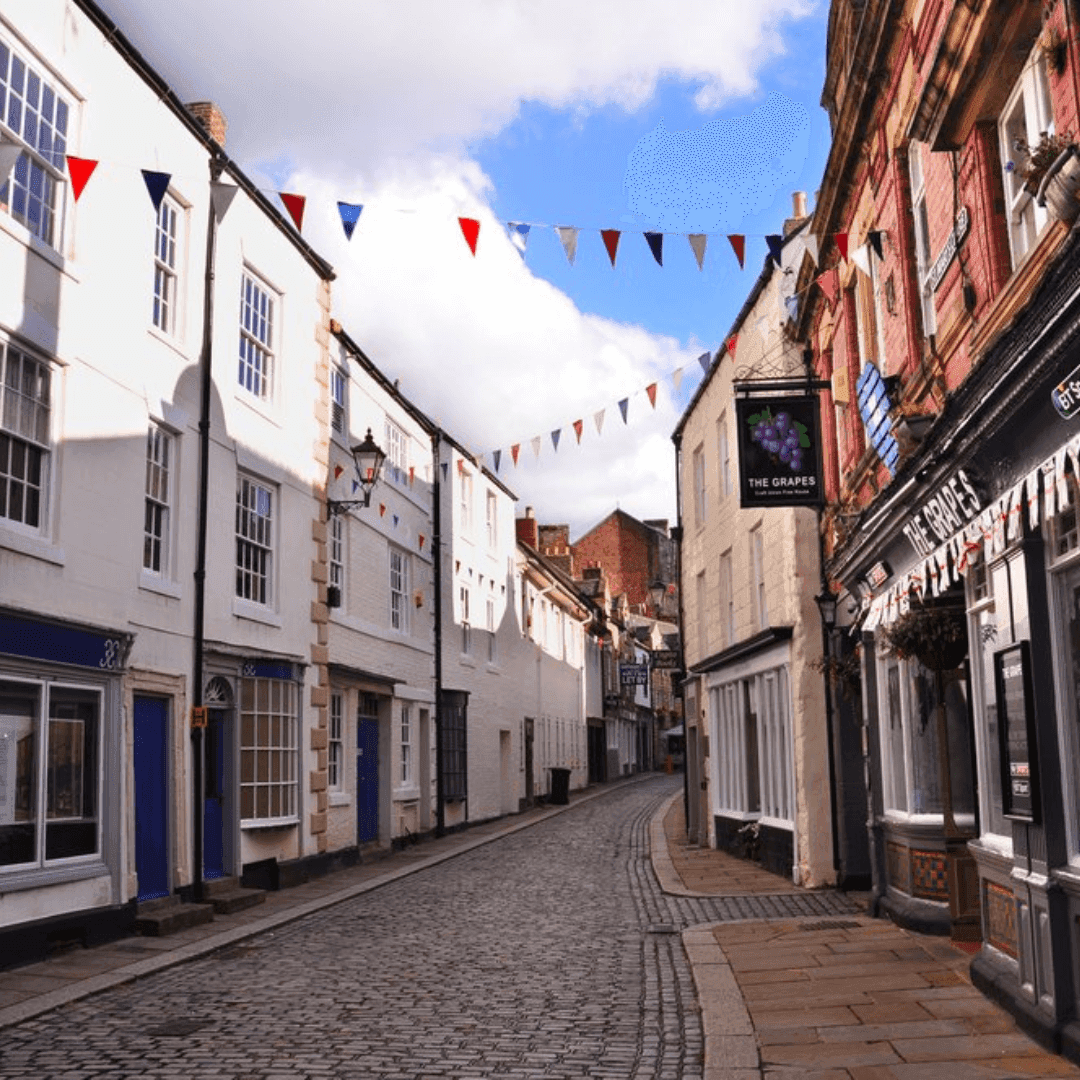A narrow cobblestone street lined with quaint shops and buildings, decorated with colorful flags in a small town. - Home Instead