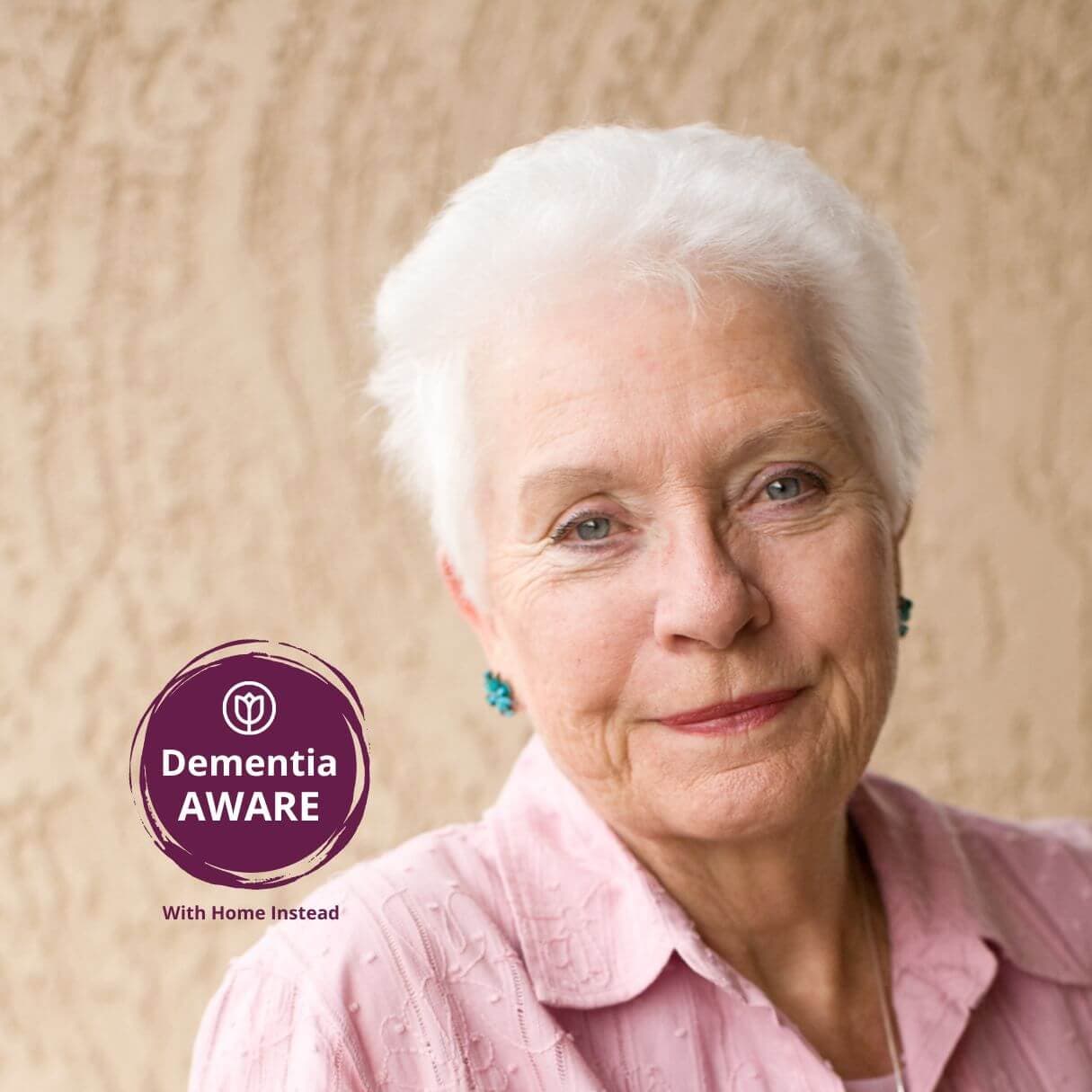 An elderly woman with short white hair wearing a pink shirt, beside a "Dementia AWARE with Home Instead" logo. - Home Instead