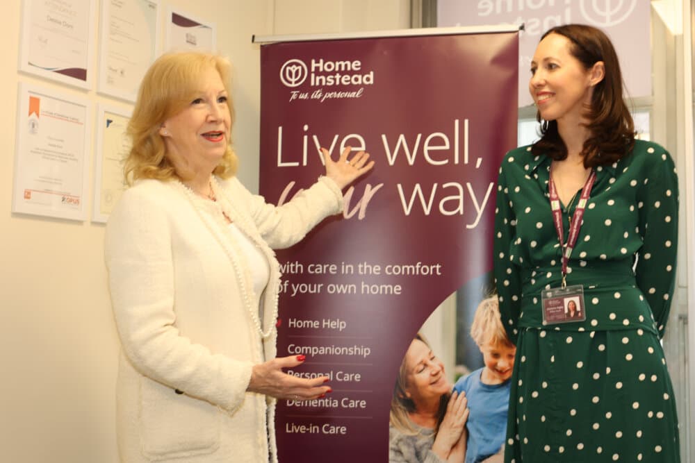 Two women stand beside a "Home Instead" banner reading "Live well, your way" promoting home care services. - Home Instead