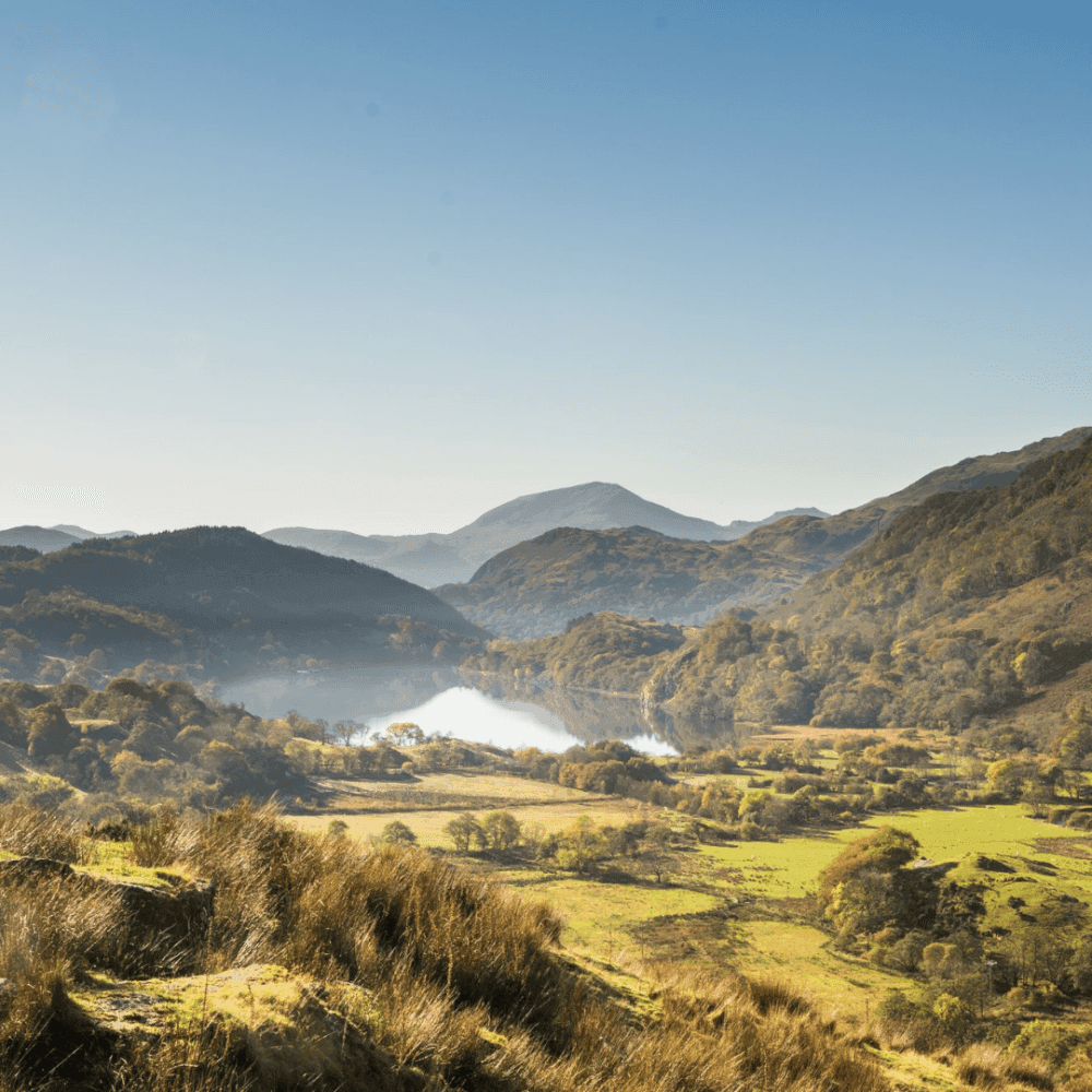 Scenic view of a sunlit valley with a misty lake, surrounded by rolling hills and mountains under a clear blue sky. - Home Instead