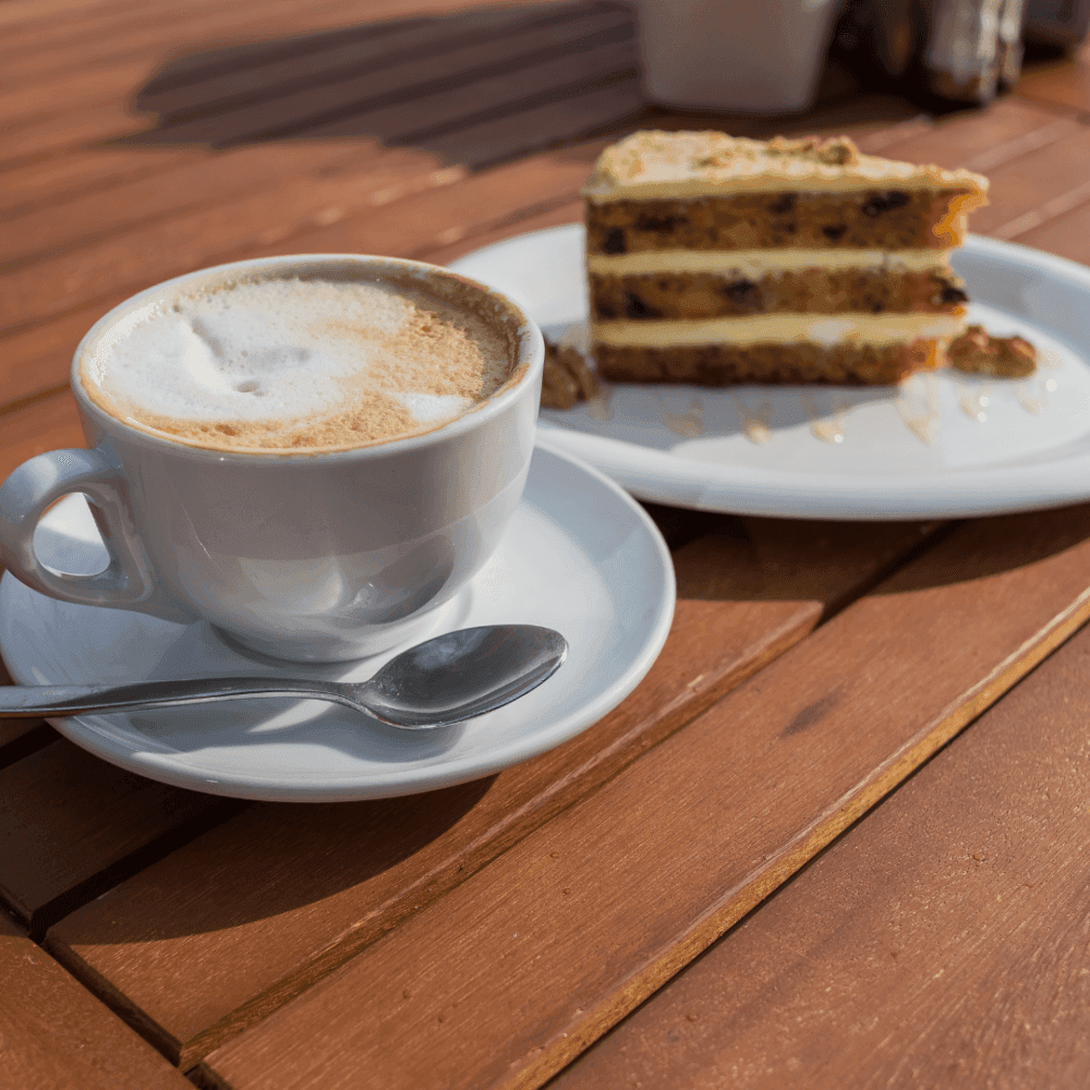 A cup of cappuccino with a spoon and a slice of layered cake on a wooden table, both served on white plates. - Home Instead