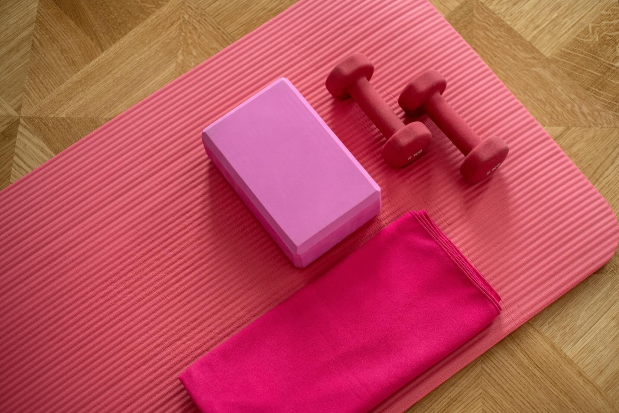 Pink yoga mat with a purple yoga block, red dumbbells, and a pink towel neatly placed on a wooden floor. - Home Instead