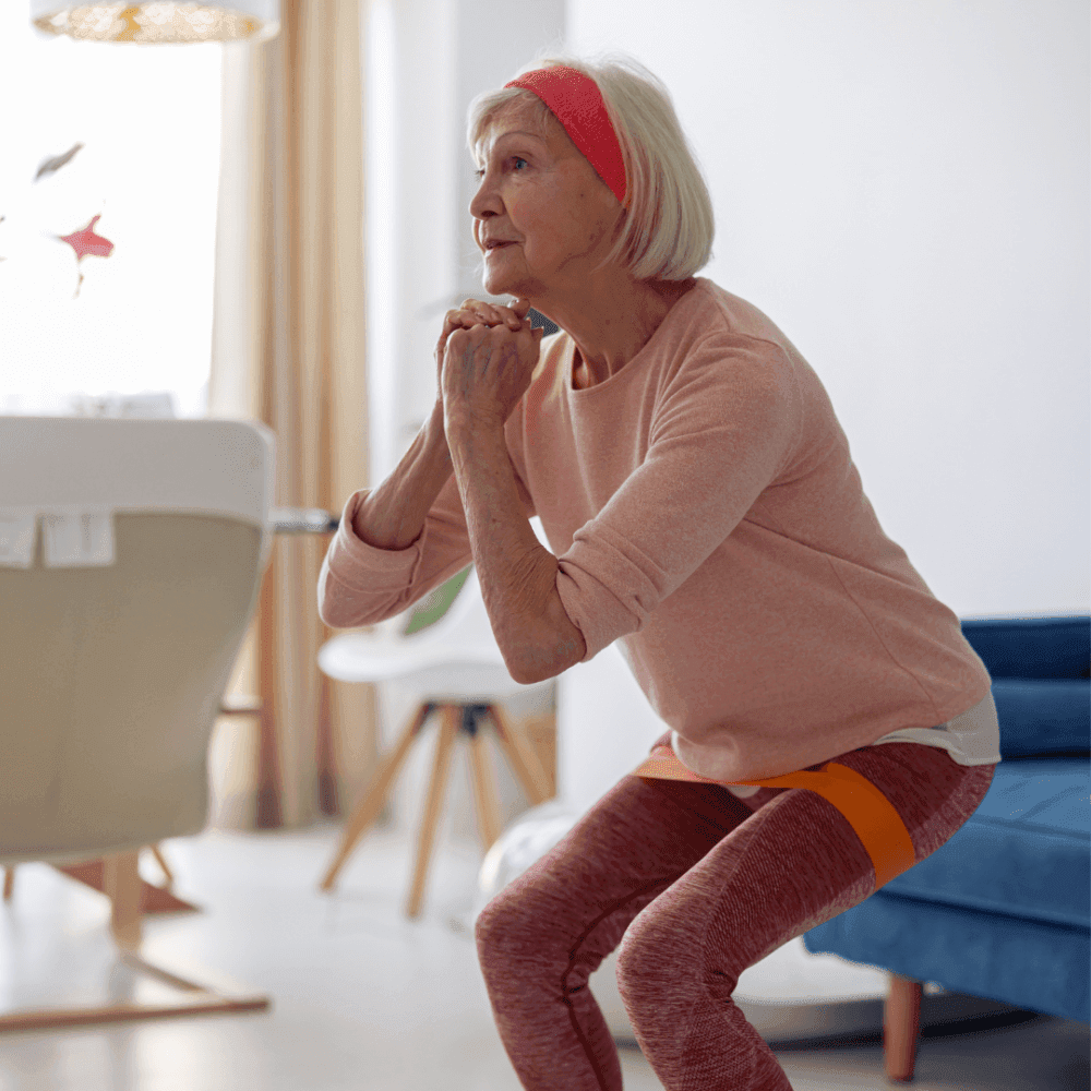 Elderly woman in athletic wear doing squats indoors with an orange resistance band around her thighs. - Home Instead