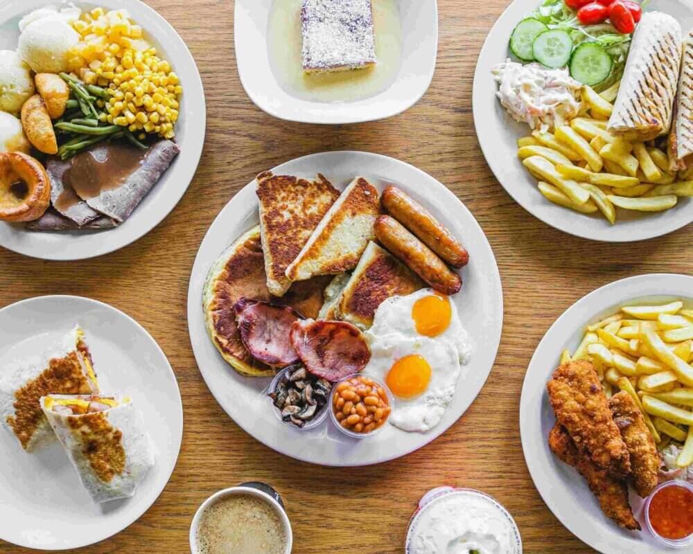 A wooden table displaying various dishes, including a full English breakfast, sandwiches, side salads, and desserts. - Home Instead