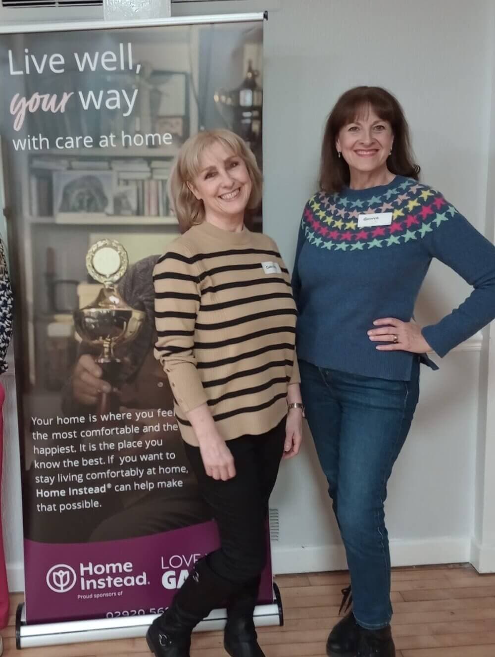 Two women smiling, standing beside a banner for Home Instead, a home care service. One wears stripes, the other a star sweater. - Home Instead