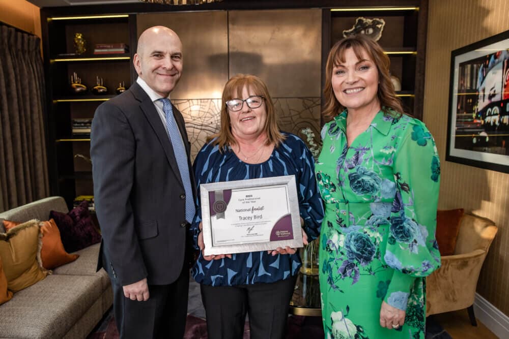 Three people, two women and one man, smile while one woman holds a framed certificate. They stand in a well-furnished room. - Home Instead