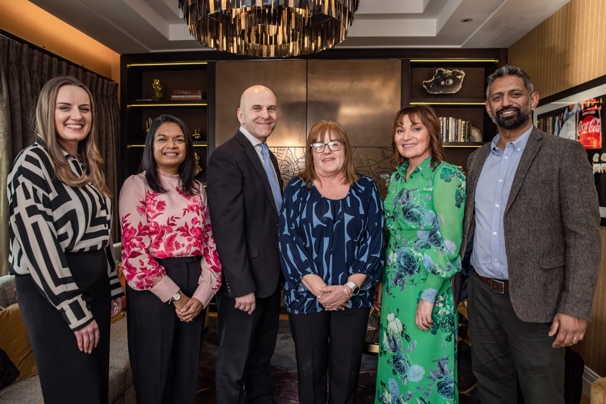 A group of six people standing together in a well-decorated room, all smiling at the camera. - Home Instead
