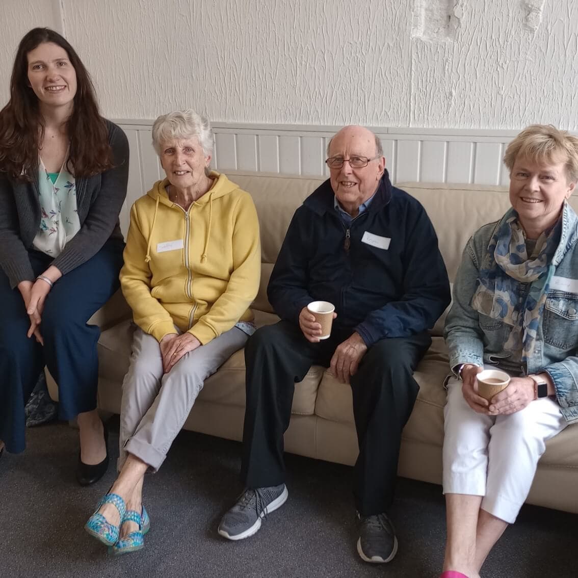 Four people are seated on a couch, smiling, some holding cups, with a light-colored wall in the background. - Home Instead
