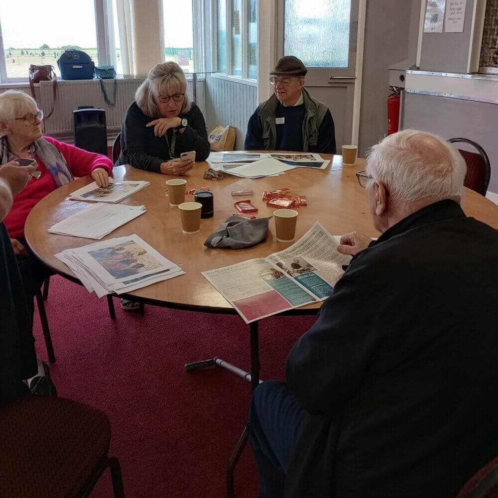 Four older adults sitting around a table, reading newspapers and chatting, with coffee cups and snacks on the table. - Home Instead