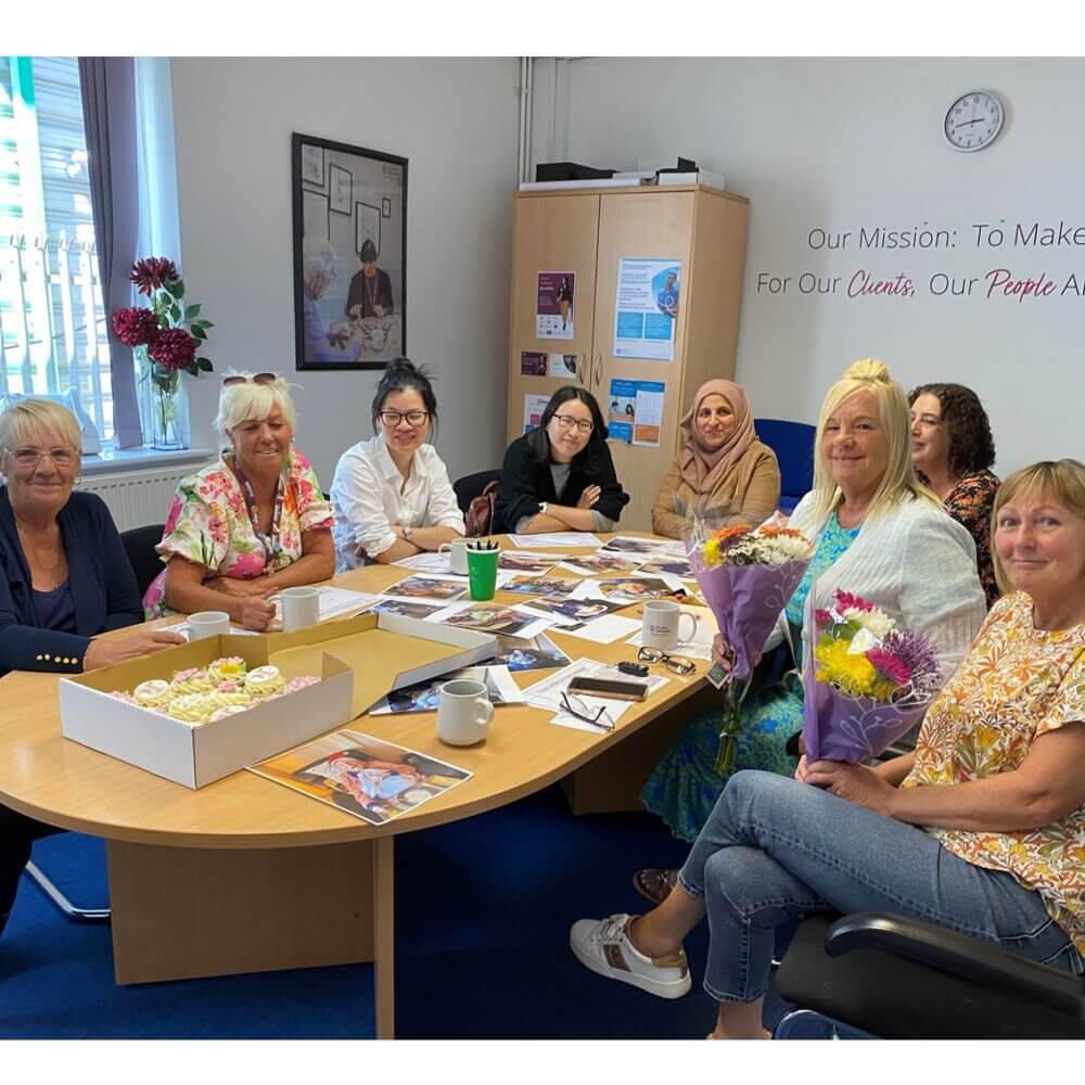 A group of eight women sit around a table with flowers, cupcakes, and documents in a brightly lit office. - Home Instead