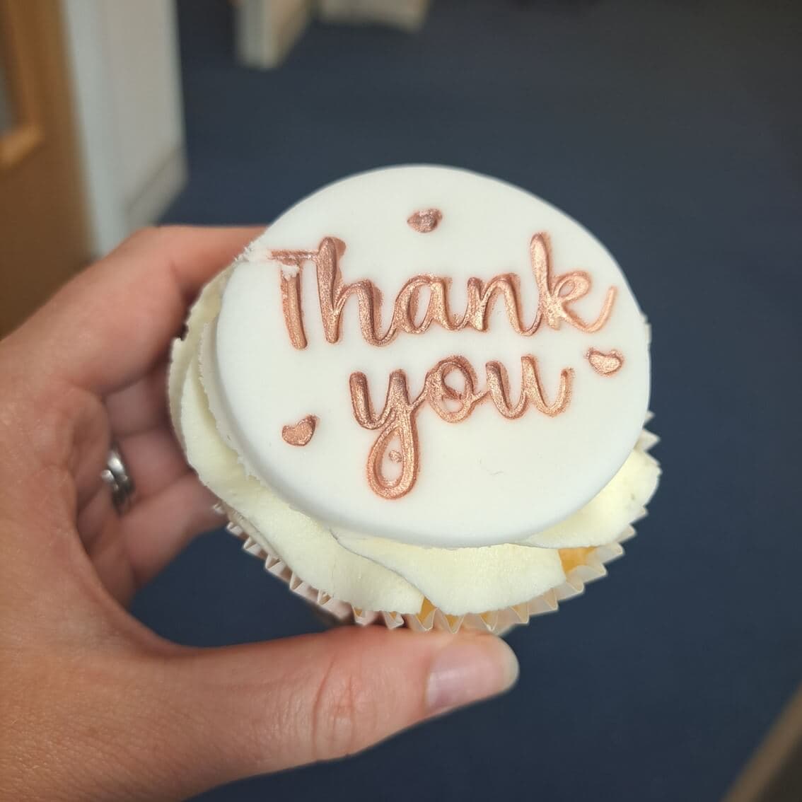 A hand holding a cupcake with white icing and "Thank you" written on top in rose gold icing. - Home Instead