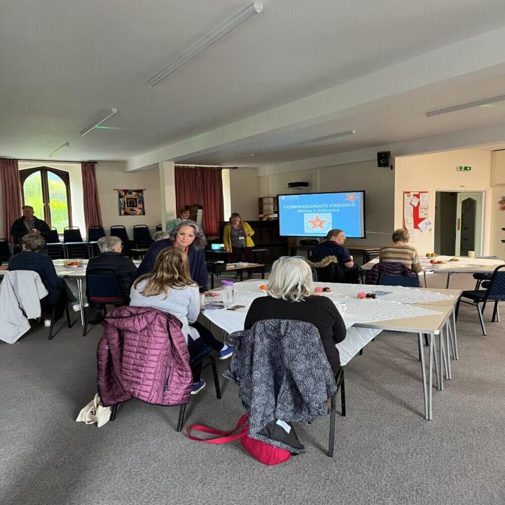 A group of people attending a meeting in a room with a presentation screen displaying "Compassionate Friends". - Home Instead