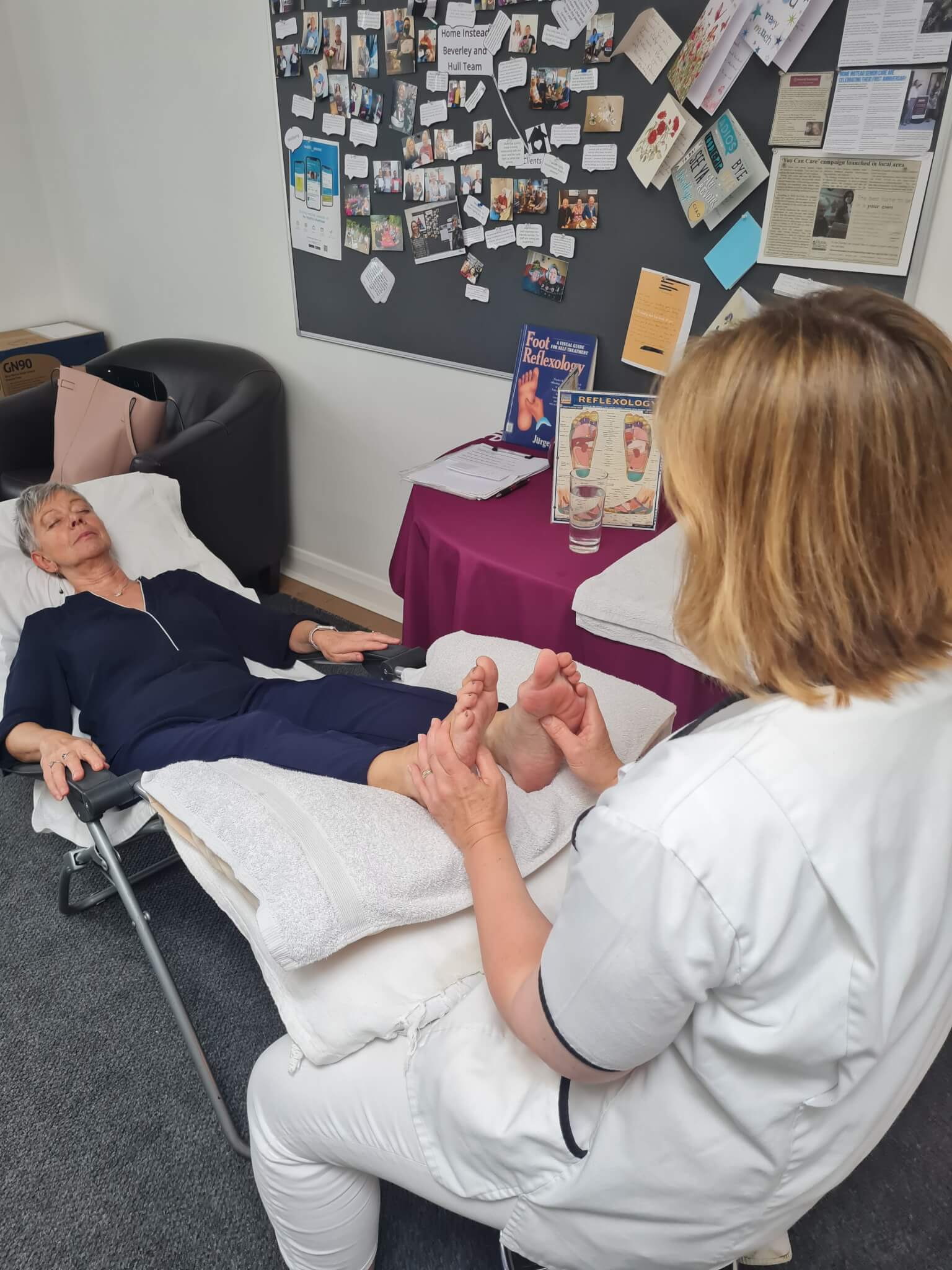 A woman receives a foot massage from a practitioner in a clinical setting. Bulletin board with various items in the background. - Home Instead