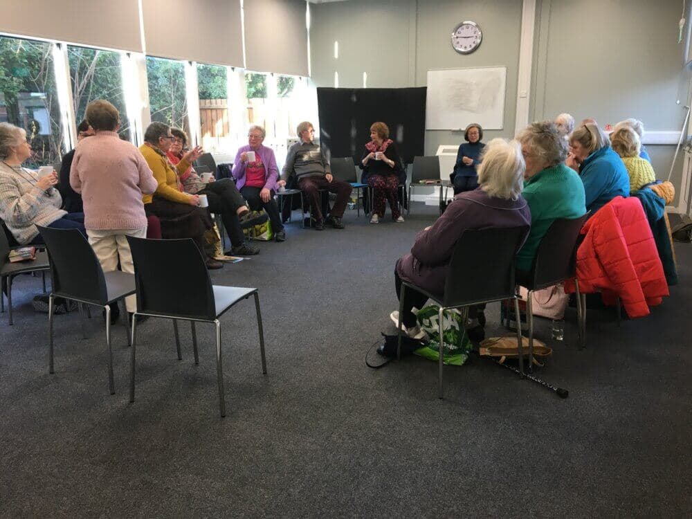 A group of elderly people sit in a circle in a room with large windows, engaged in a discussion or activity. - Home Instead