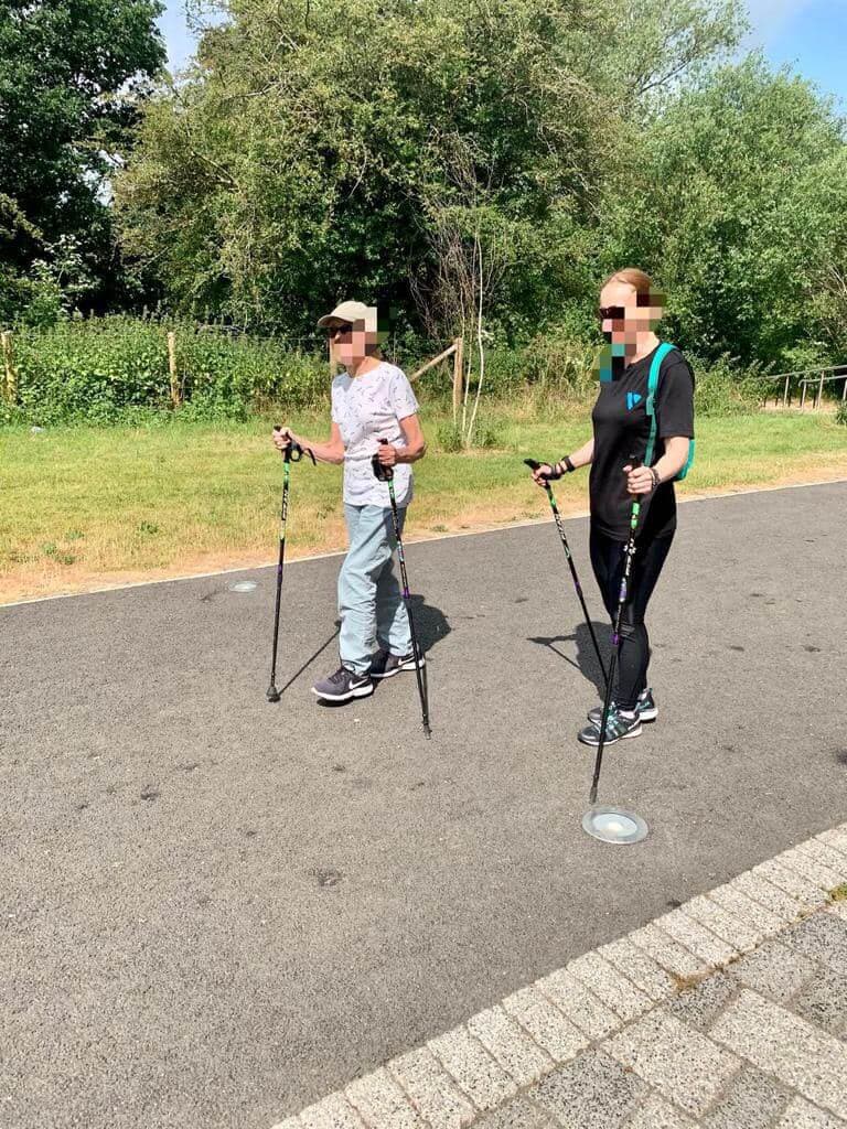 Two people are Nordic walking on a paved path in a park, with trees and grass in the background. Their faces are pixelated. - Home Instead