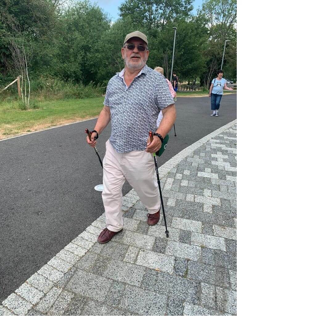 An elderly man walking with trekking poles on a paved path, wearing a hat, sunglasses, a patterned shirt, and light pants. - Home Instead