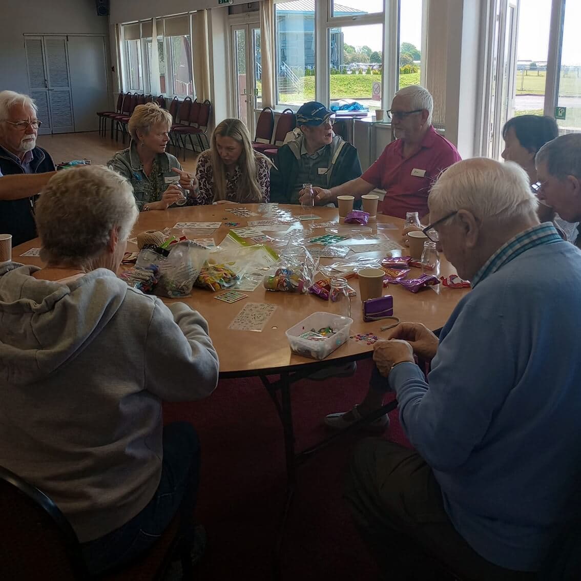 A group of people sitting around a table, working on crafts in a well-lit room with large windows. - Home Instead