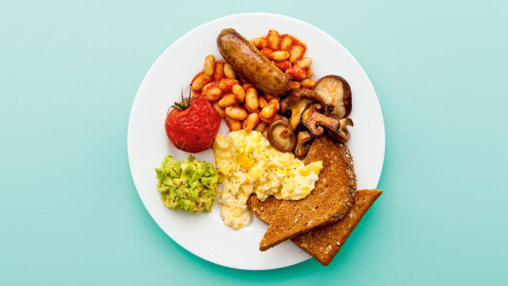 A plate with scrambled eggs, avocado, grilled tomato, sausage, baked beans, toast, and mushrooms on a blue background. - Home Instead