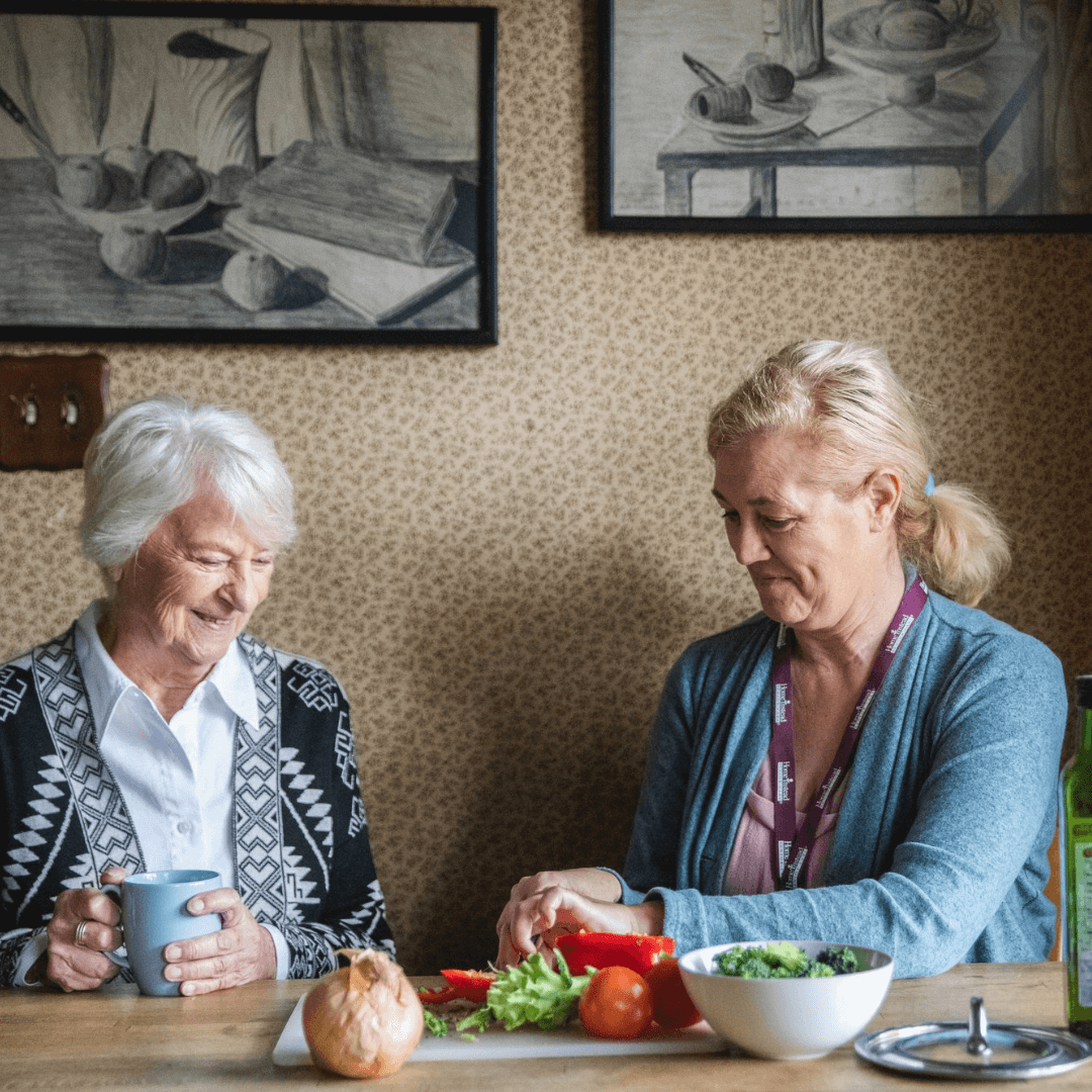 Two women in a kitchen; one is chopping vegetables while the other holds a mug and smiles. - Home Instead