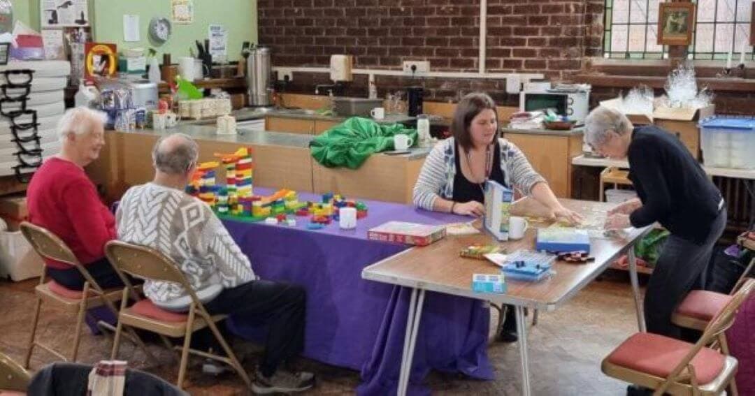 Four elderly individuals engage in activities at tables in a community room with brick walls and kitchen appliances. - Home Instead