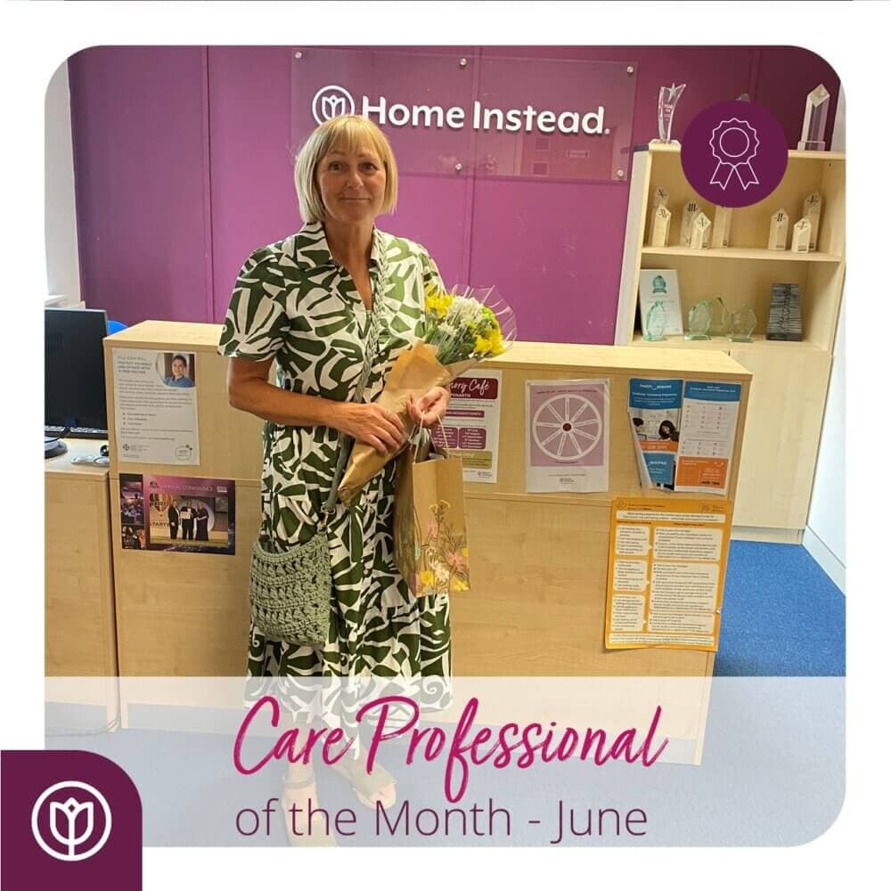 A person stands smiling with flowers and a gift in an office. Text reads "Care Professional of the Month - June. - Home Instead