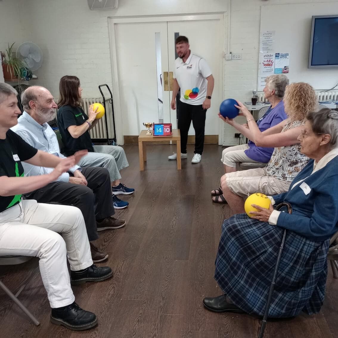 A group of elderly people sit in chairs, playing a ball game, while a man stands and supervises near a score board. - Home Instead