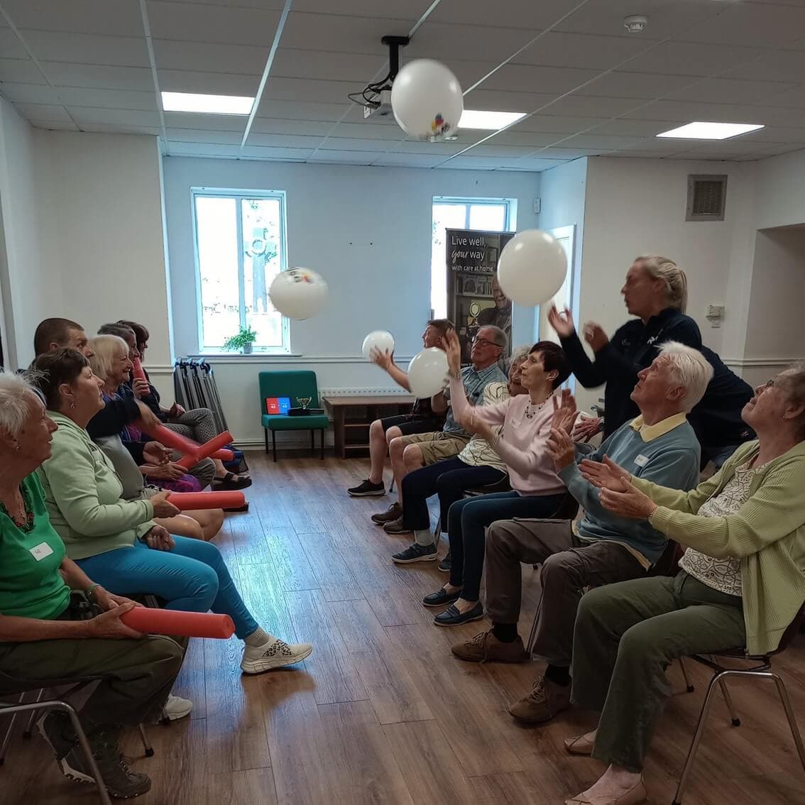 A group of elderly people sitting in chairs in a circle, joyfully playing with white balloons indoors. - Home Instead