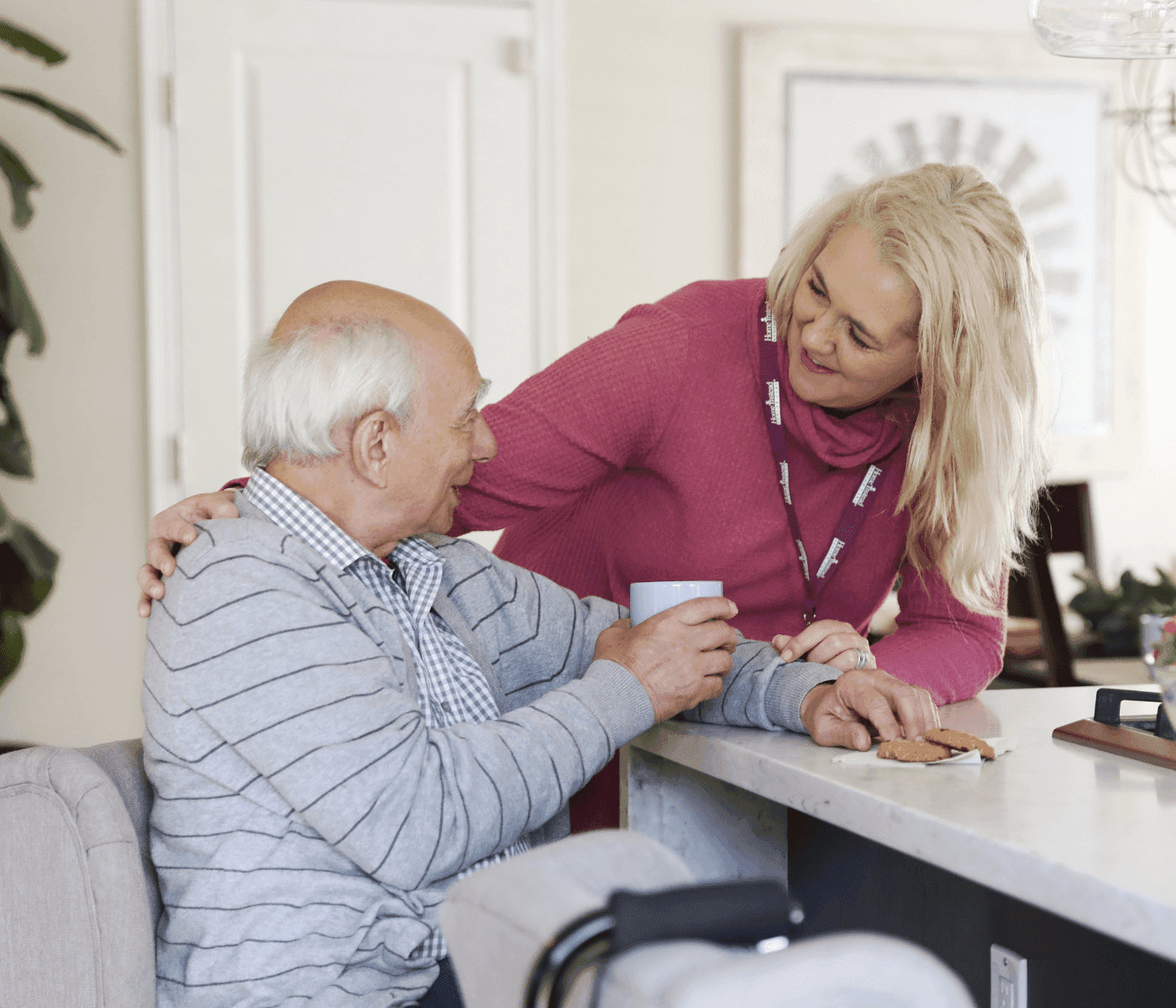 A caregiver with a name badge smiles and comforts an elderly man holding a cup at a kitchen counter. - Home Instead