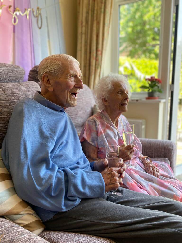 An elderly couple sits happily on a couch, holding glasses of champagne and smiling in a brightly lit room. - Home Instead