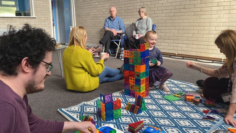 Adults and children playing with colorful building blocks on a carpet in a room with brick walls. - Home Instead