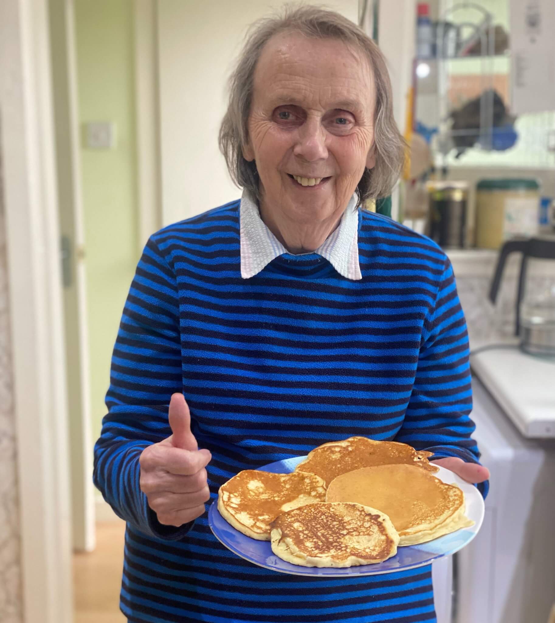 Older adult in a striped sweater smiles, holding a plate of pancakes and giving a thumbs-up in a kitchen. - Home Instead