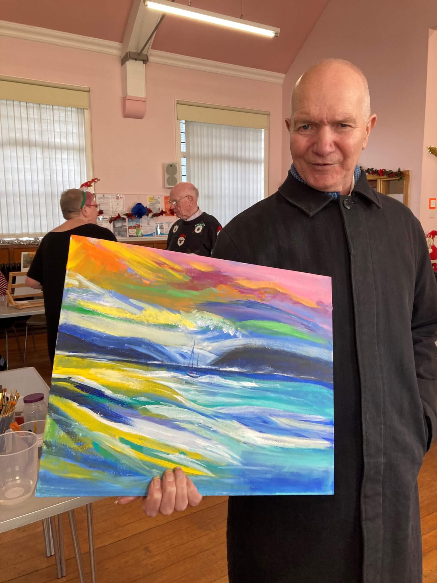 A man holding a colorful abstract painting in an art room with people and art supplies in the background. - Home Instead