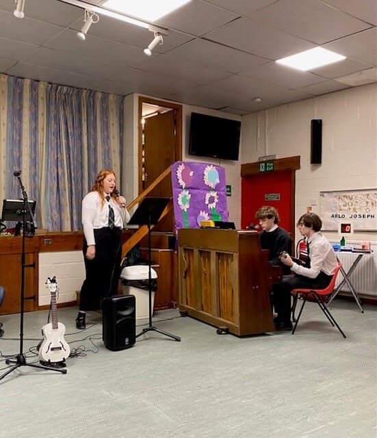 Three people performing music indoors: one singing, one playing piano, and another seated. A guitar is also visible. - Home Instead