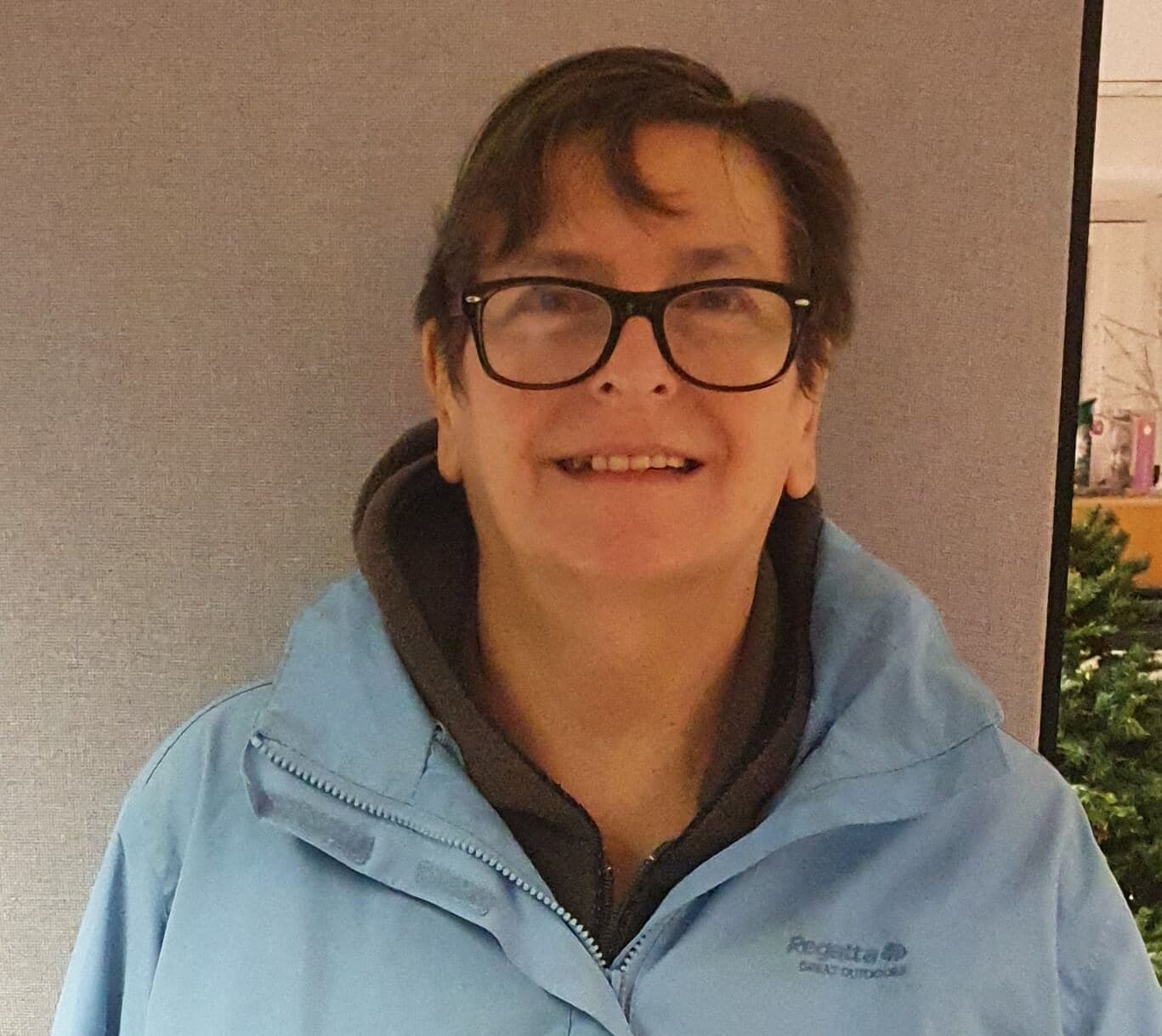 A person with short hair and glasses smiles while wearing a blue jacket in front of a gray background. - Home Instead