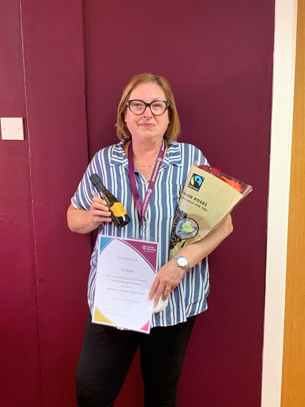 A woman with glasses holds a certificate, wine bottle, and bag of Fairtrade roasted coffee in front of a maroon wall. - Home Instead