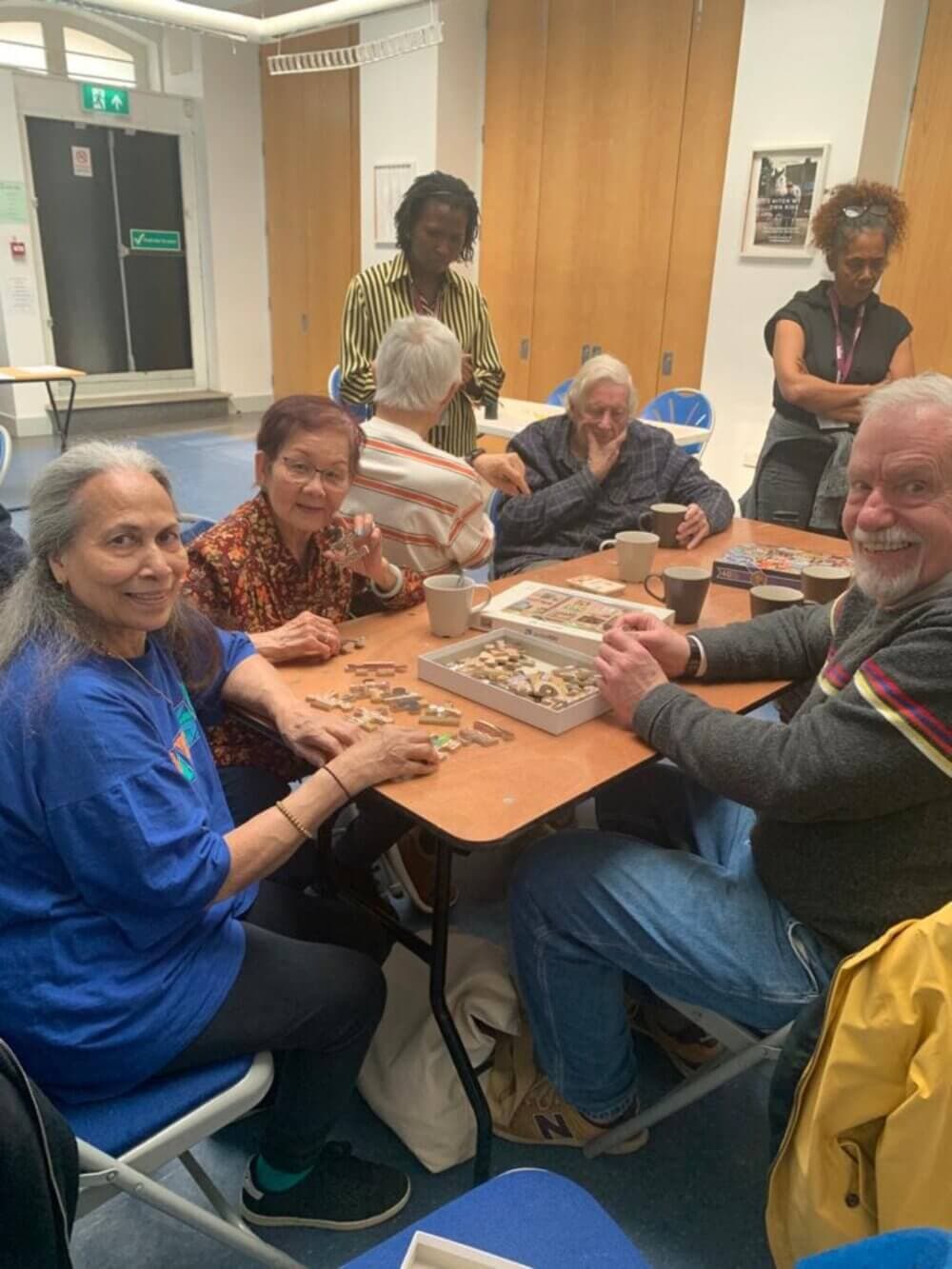 A group of people, young and old, sit around a table working on a jigsaw puzzle, smiling and engaging with each other. - Home Instead