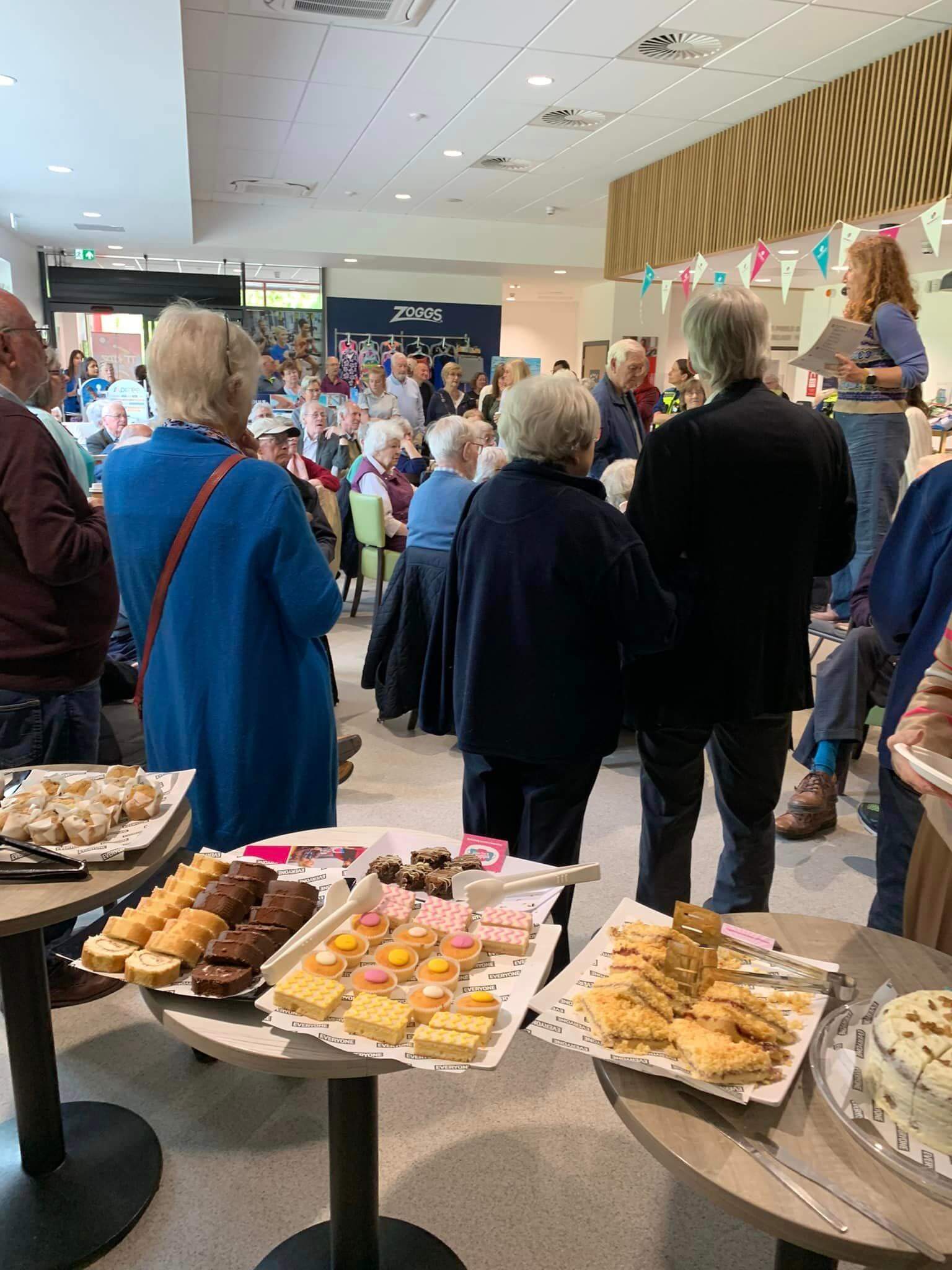 A group of people at an indoor event with tables of assorted pastries and desserts in the foreground. - Home Instead