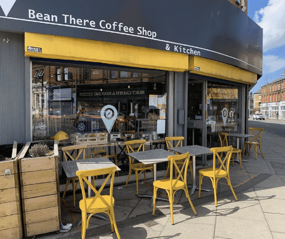 Outdoor seating area at "Bean There Coffee Shop & Kitchen" with yellow chairs and tables facing a glass storefront. - Home Instead