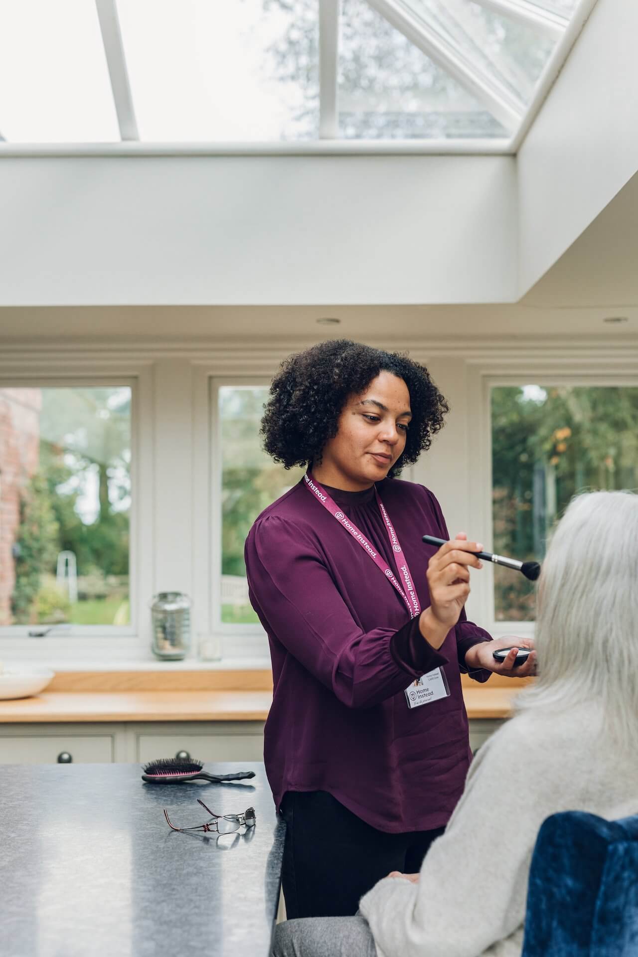 A caregiver applies makeup to an elderly person sitting in a brightly lit room with large windows. - Home Instead