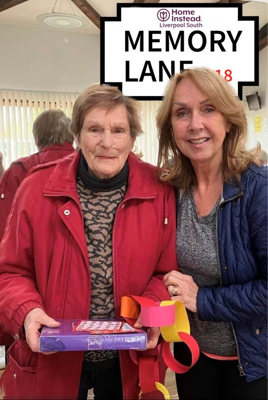 Two women smiling in front of a "Memory Lane 2018" sign. One holds a gift and colorful paper chain. - Home Instead