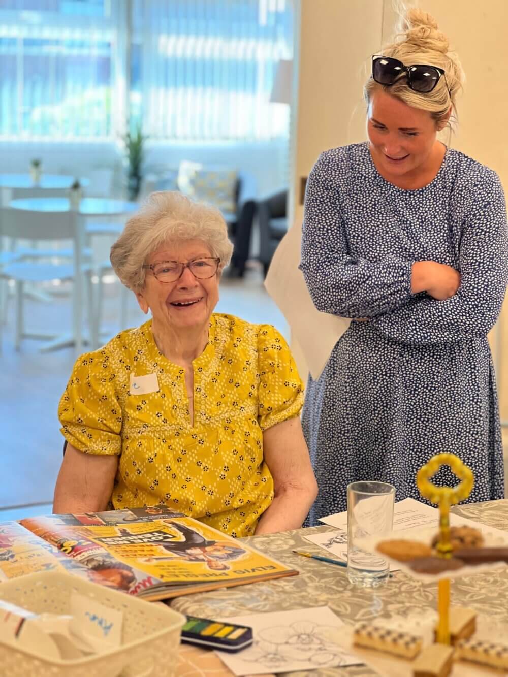 An elderly woman in a yellow blouse smiles while a younger woman in a blue dress stands beside her, both in a bright room. - Home Instead