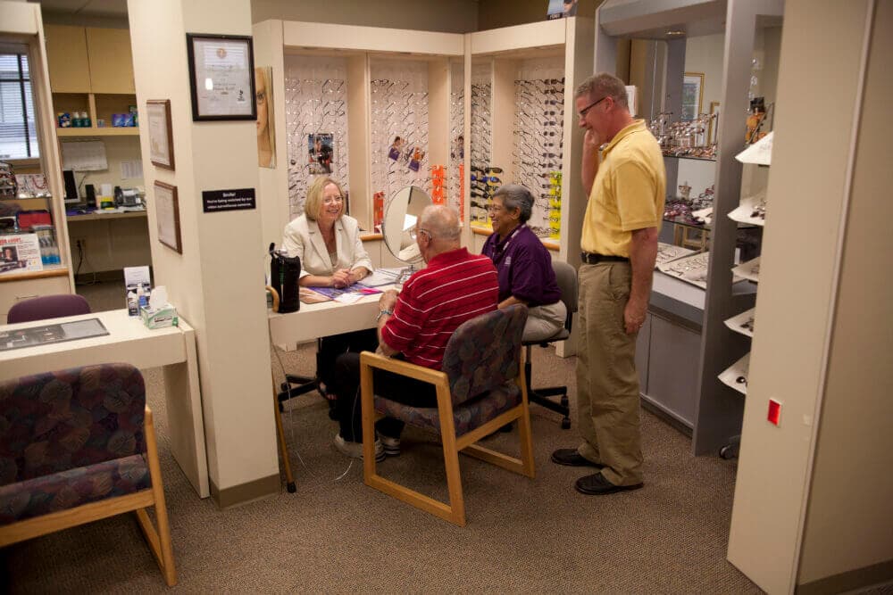 Four people in a small optician's office, sitting and standing near a desk, surrounded by eyeglasses displays. - Home Instead