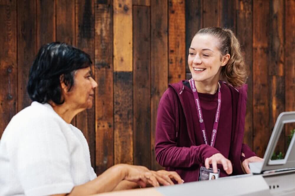 A woman helping another woman use a laptop, both smiling and seated at a wooden table. - Home Instead